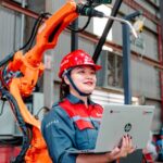 Female engineer with laptop overseeing robotic arm in a manufacturing setting in Vietnam.