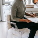 From above of unrecognizable female psychologist with crossed legs taking notes on clipboard while filling out questionnaire during psychotherapy appointment in cozy office