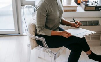 From above of unrecognizable female psychologist with crossed legs taking notes on clipboard while filling out questionnaire during psychotherapy appointment in cozy office