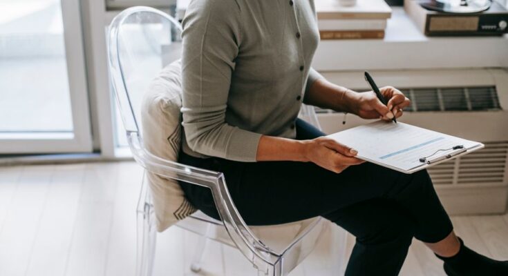 From above of unrecognizable female psychologist with crossed legs taking notes on clipboard while filling out questionnaire during psychotherapy appointment in cozy office