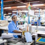 A female factory worker in India manages a sewing machine in a bustling textile factory.