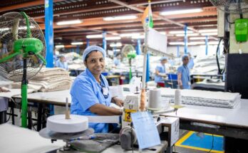 A female factory worker in India manages a sewing machine in a bustling textile factory.