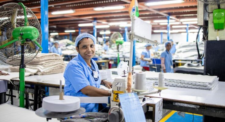 A female factory worker in India manages a sewing machine in a bustling textile factory.